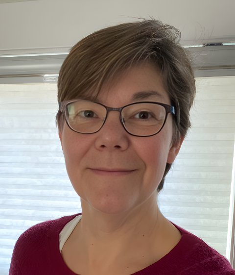 A woman with short brown hair and glasses smiling indoors near a window with white blinds.
