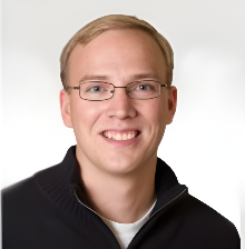 Portrait of a young man with blonde hair, glasses, and a black zip-up sweater smiling against a white background.