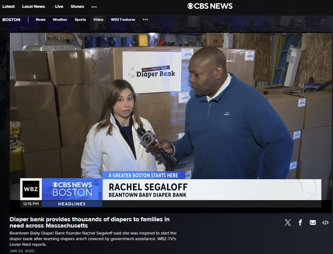 Rachel Segaloff, founder of Beantown Baby Diaper Bank, is being interviewed by a male reporter holding a microphone at a warehouse with stacks of cardboard boxes labeled for diaper distribution.