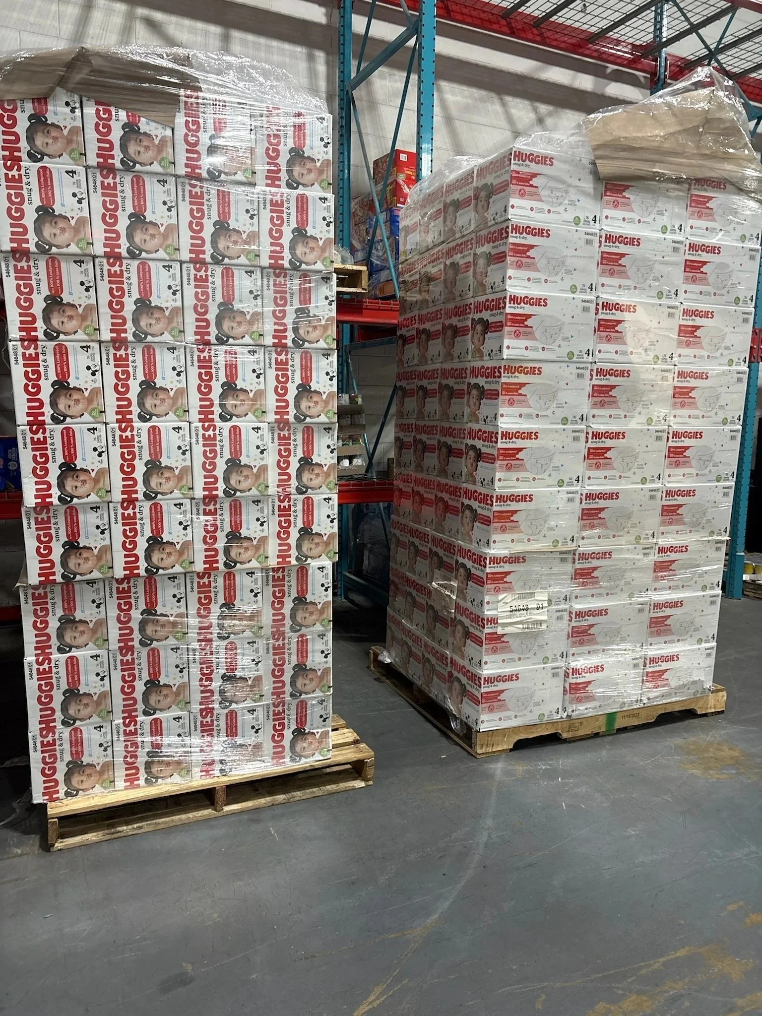 Stacks of boxes of Huggies baby diapers on pallets in a warehouse store aisle.
