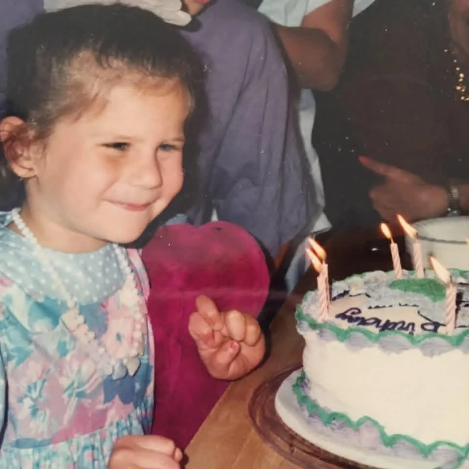 A young girl celebrating her birthday with a cake on a table, with lit candles on top.