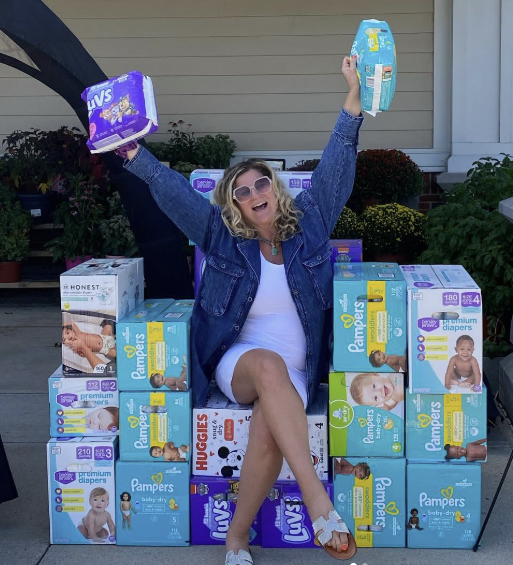 Woman sitting on a pile of diaper boxes, holding diapers in each hand, smiling and celebrating outdoors.