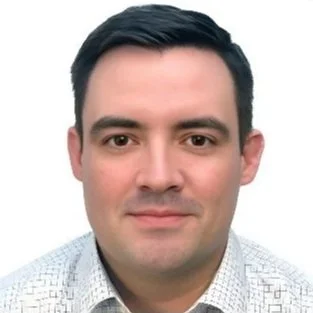 Professional headshot of a man with dark hair wearing a light-colored, patterned shirt against a plain background.