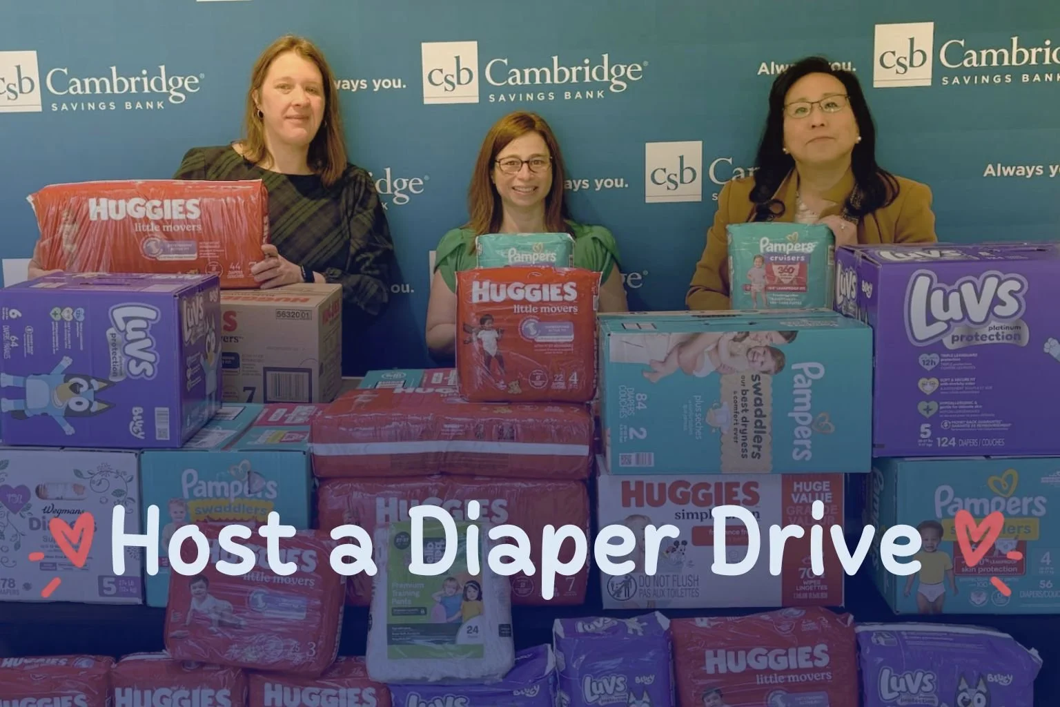 Three women stand behind stacks of donated diapers during a Cambridge Savings Bank diaper drive.