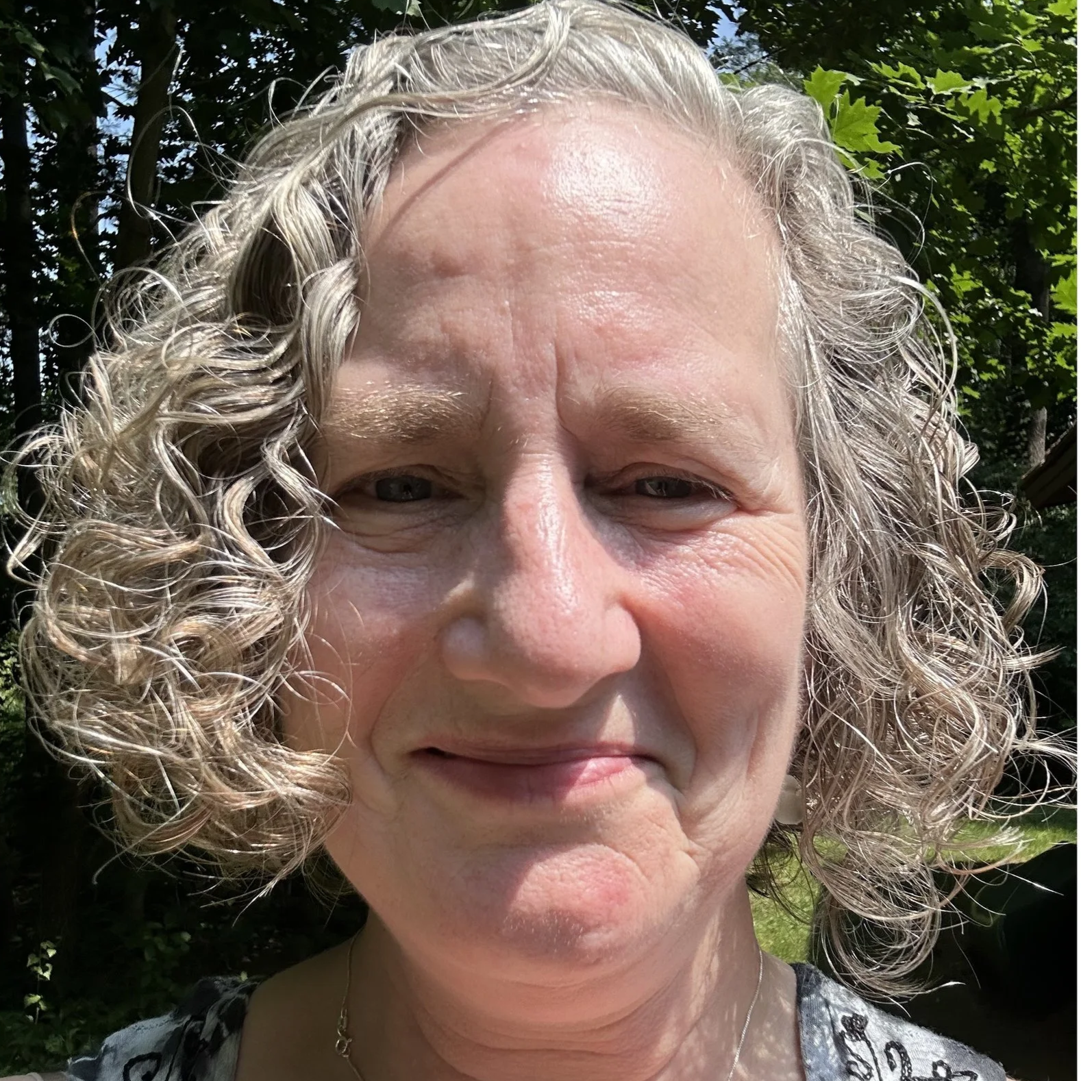 A close-up of a woman with curly, gray hair outdoors, smiling slightly, with a background of trees and foliage.