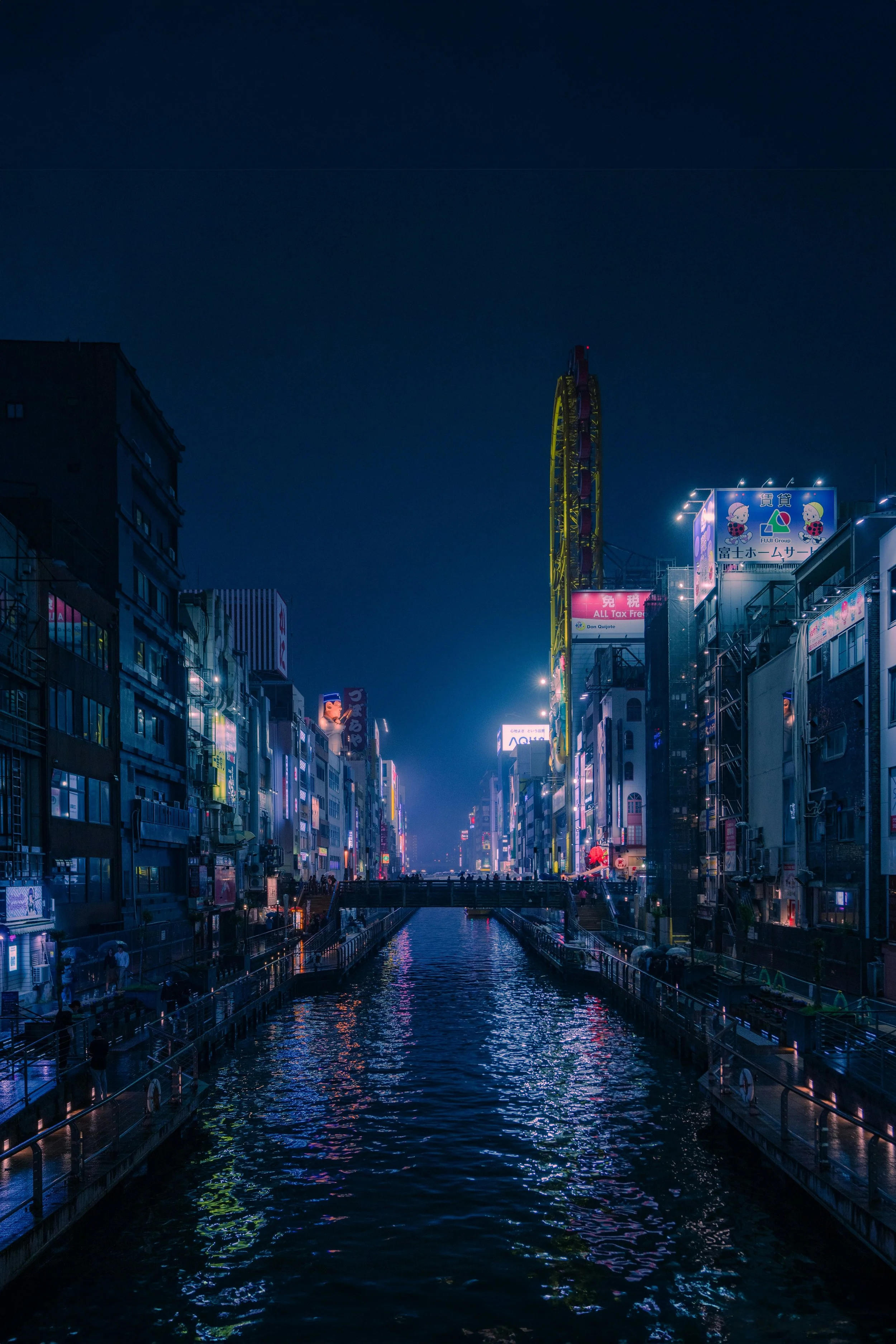 Night scene of a city with illuminated signs and buildings lining a canal, reflecting colorful lights on the water.