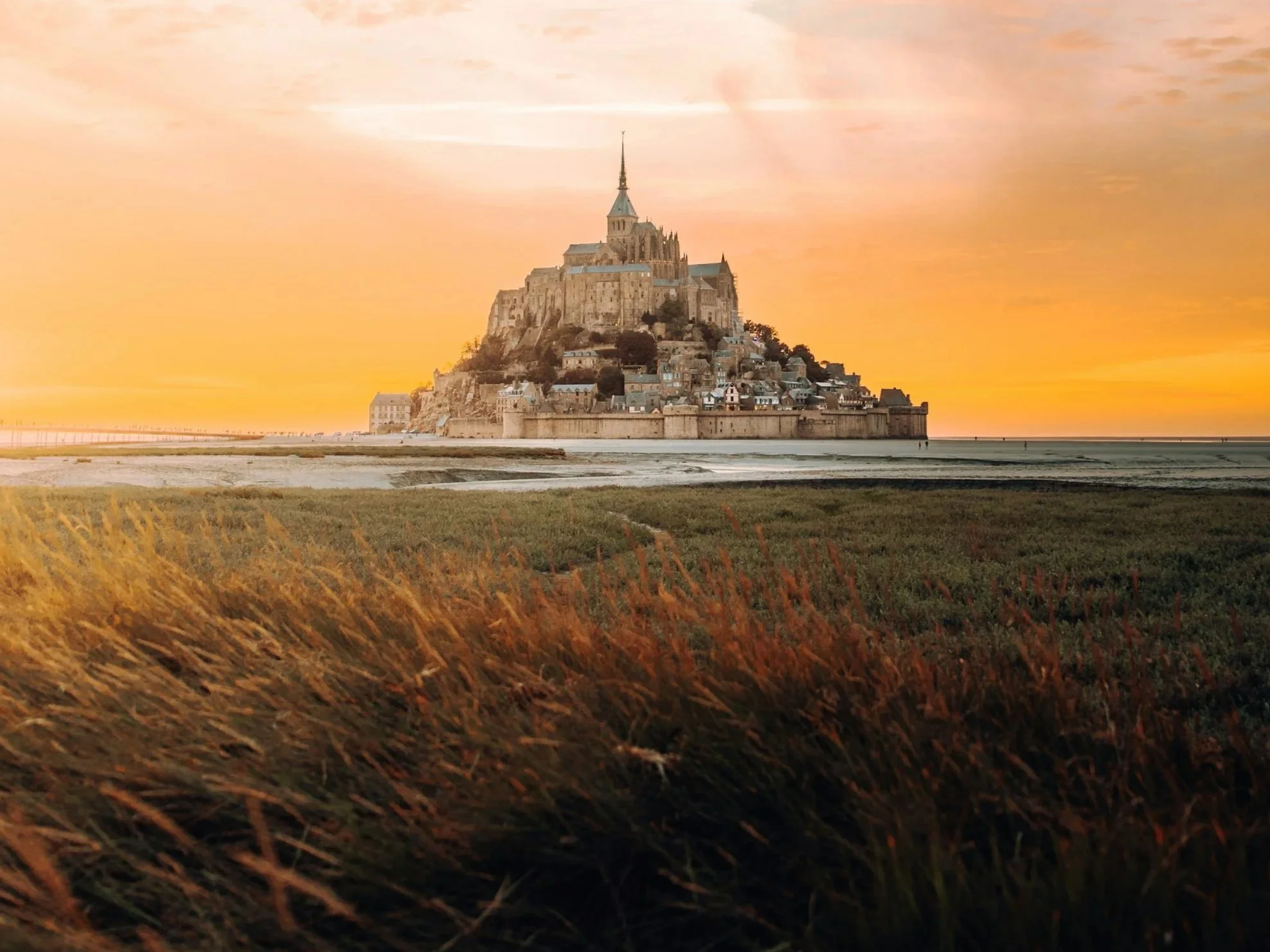 View of Mont Saint-Michel during sunset with sky painted in warm orange and pink hues, surrounded by water and grassy marshland.