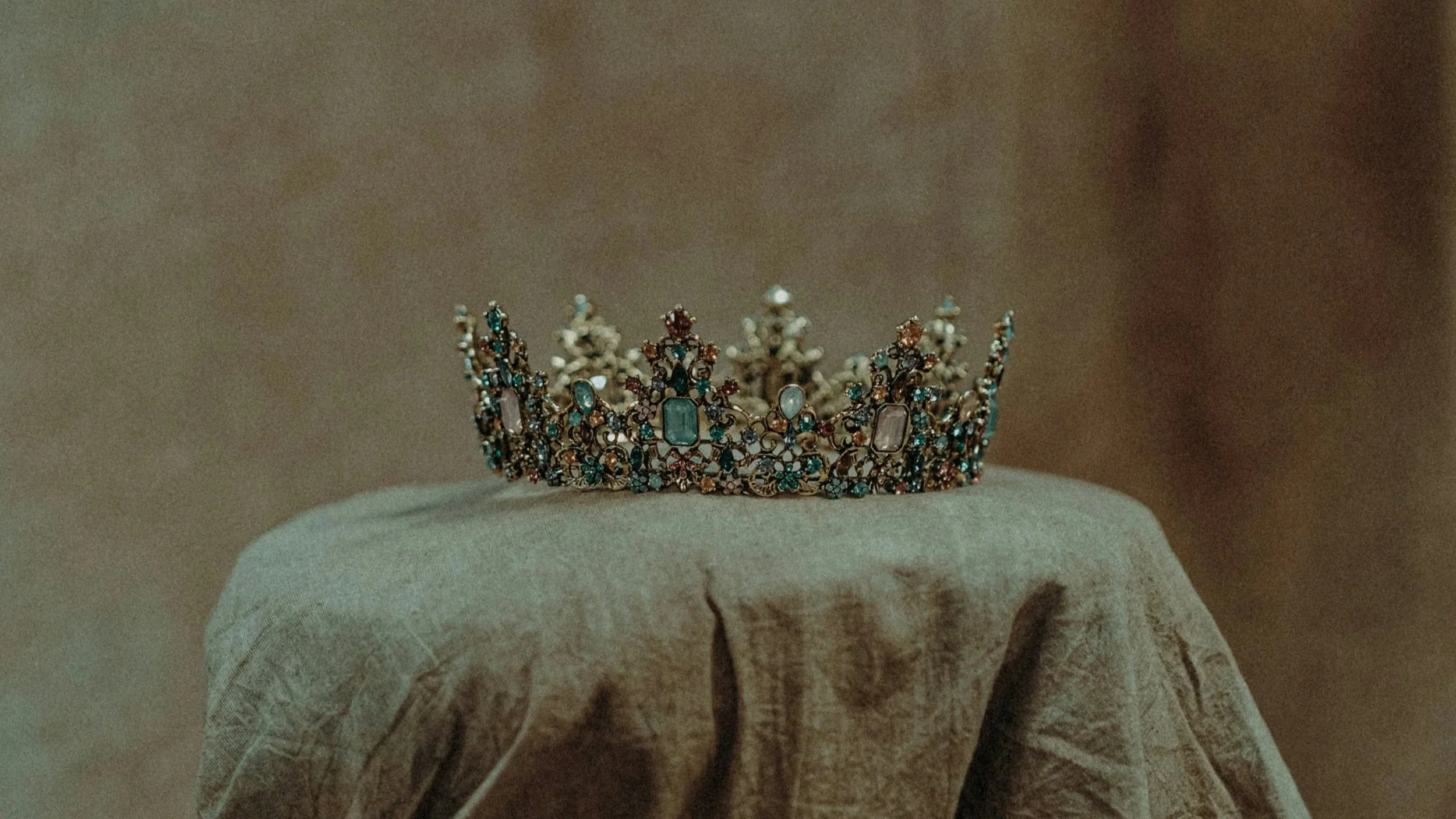 A jeweled crown with various gemstones displayed on a pedestal covered by a beige cloth.