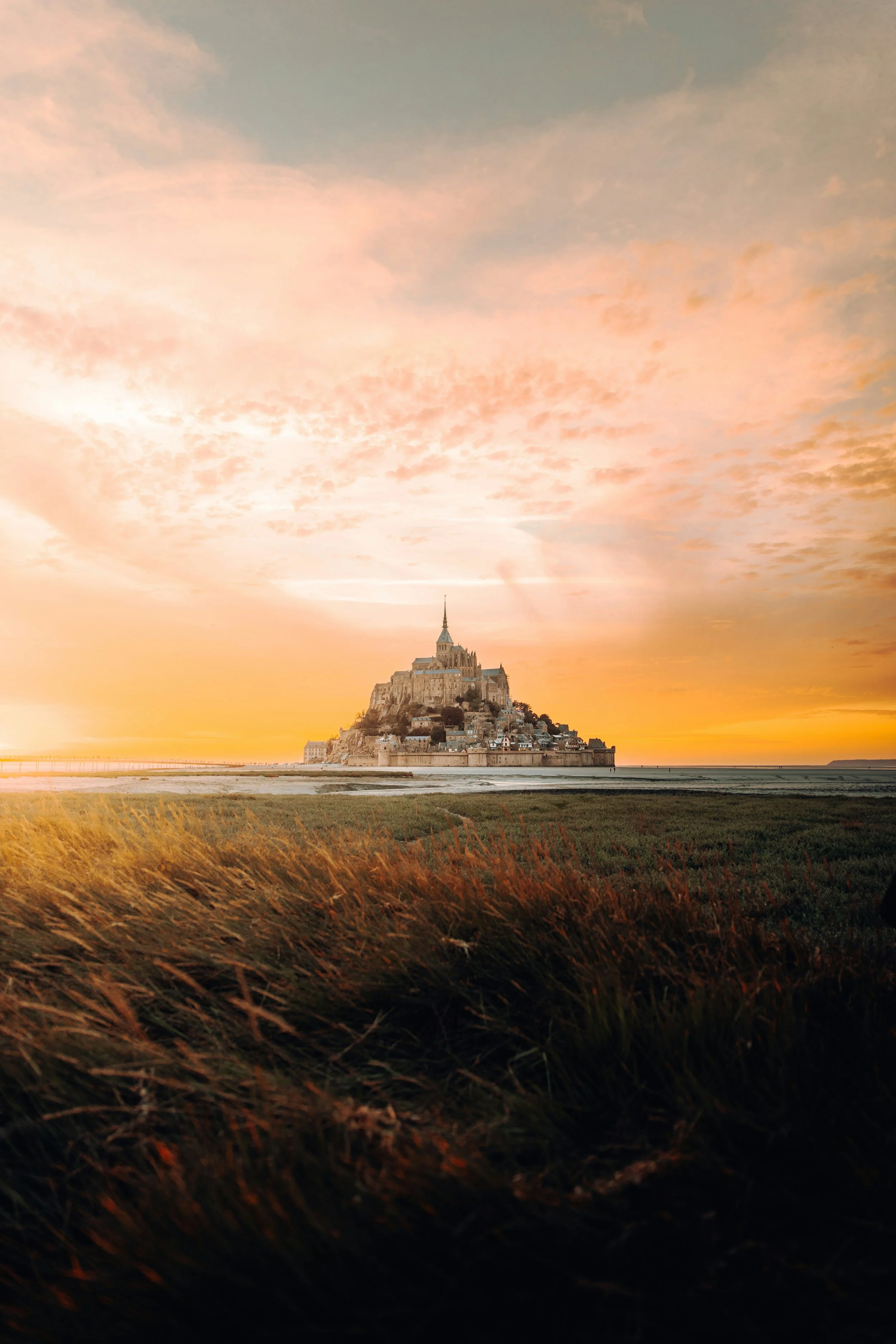 Sunset at Mont Saint-Michel, a historic island commune in France, with a cloudy sky and warm glow, viewed from the seaside with grassy foreground.