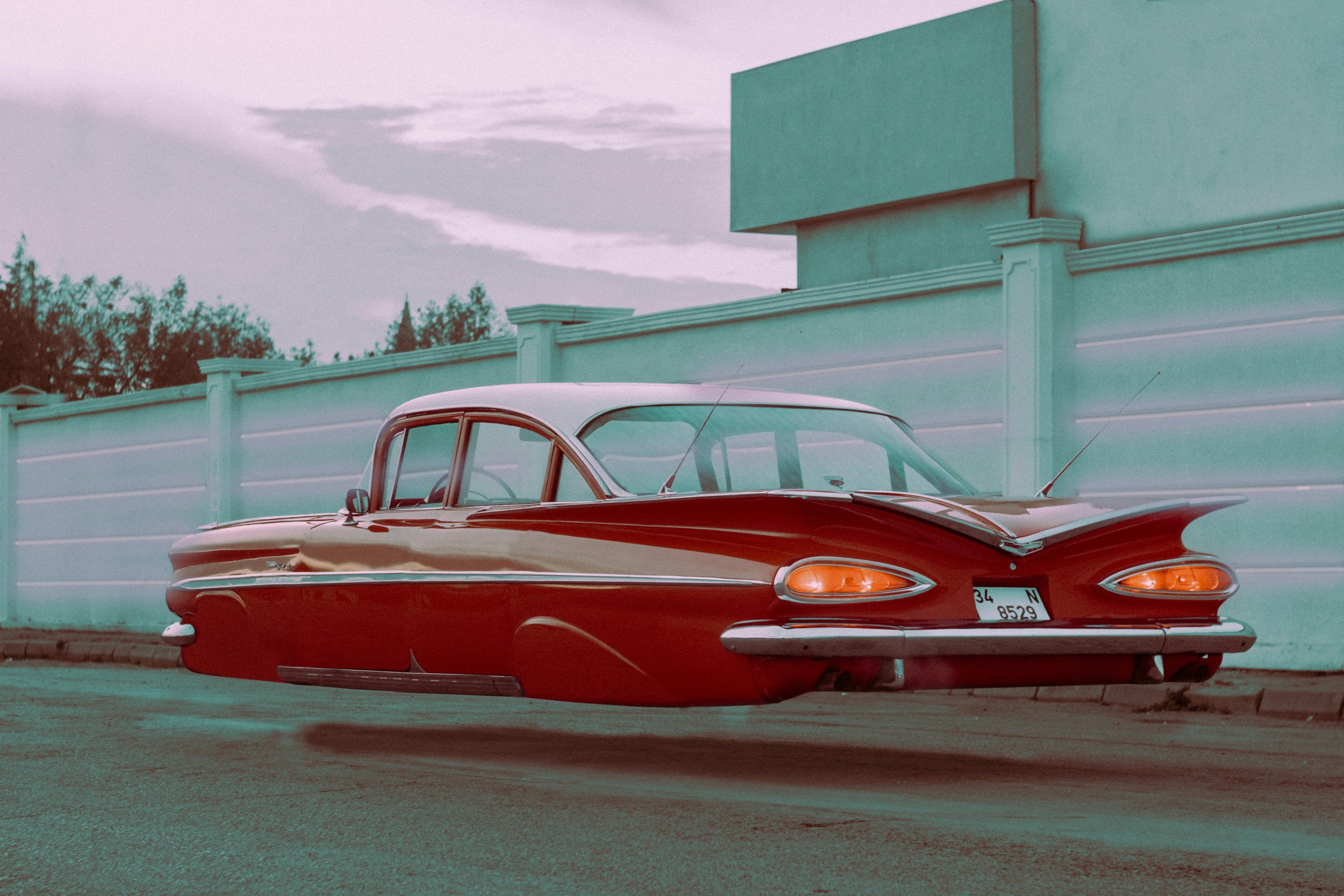A vintage red car parked next to a white fence, with trees and a cloudy sky in the background.