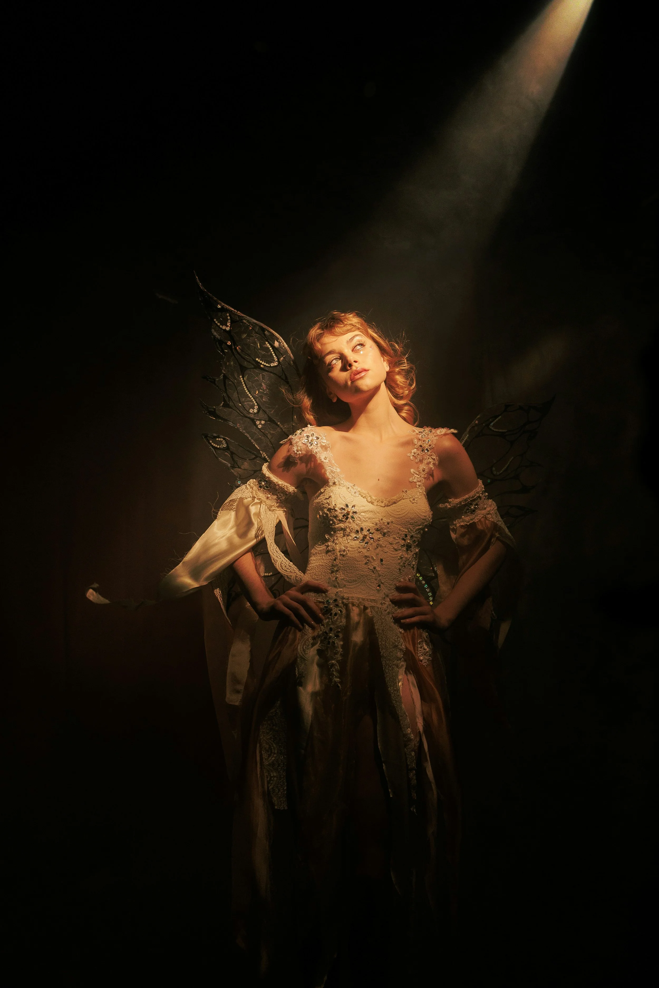 A woman dressed as a fairy with fairy wings, elegant gown, and styled hair, standing on stage under a spotlight.