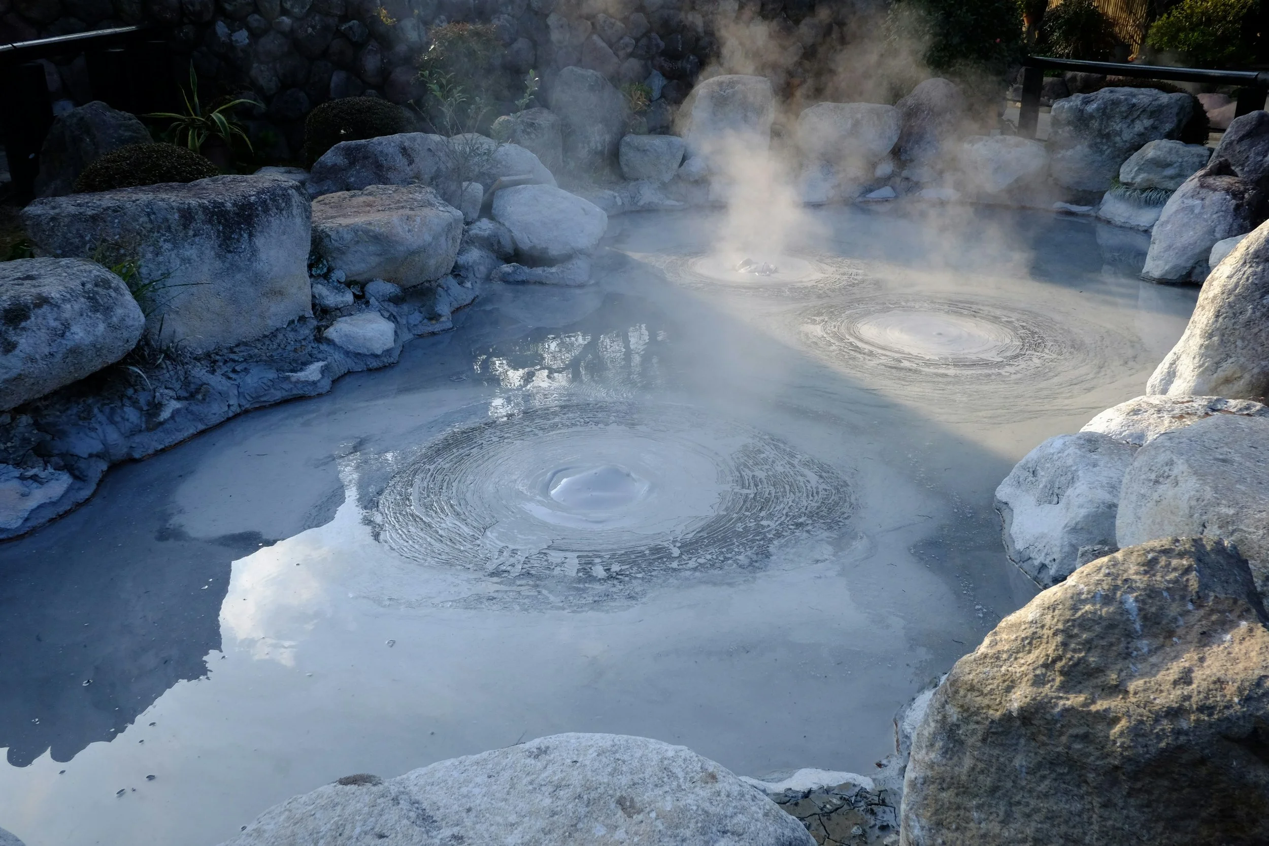 A hot spring with steaming water surrounded by large rocks and stones, with some plants and a black railing visible in the background.
