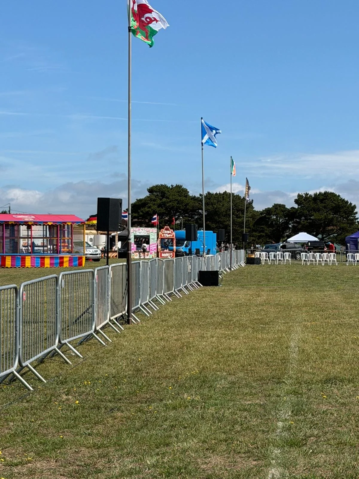 An outdoor fair or festival with food stalls, flags, chairs, barriers, and a clear blue sky.