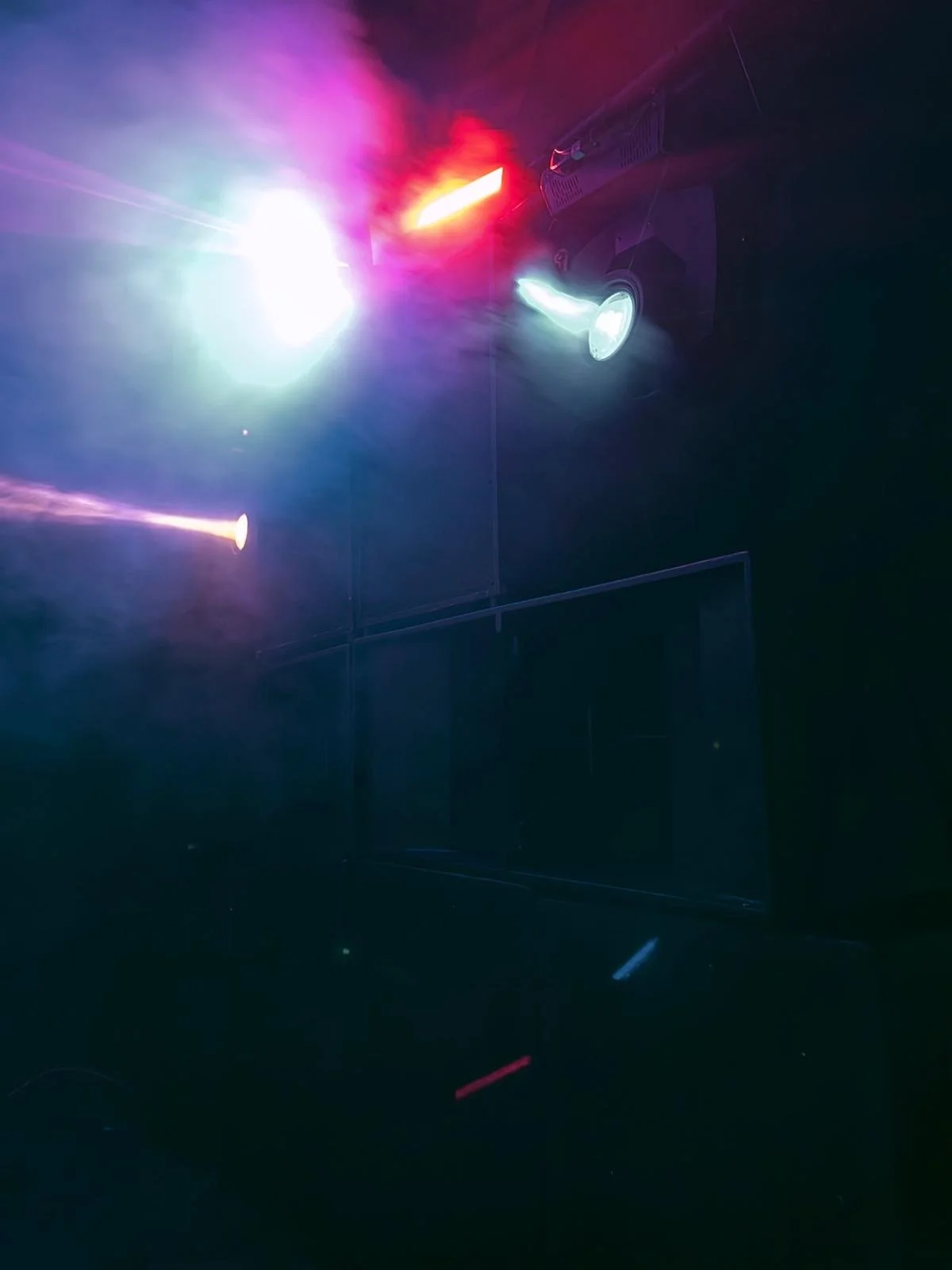 Dark room with bright, colorful stage lights in red, green, and purple creating a hazy atmosphere.