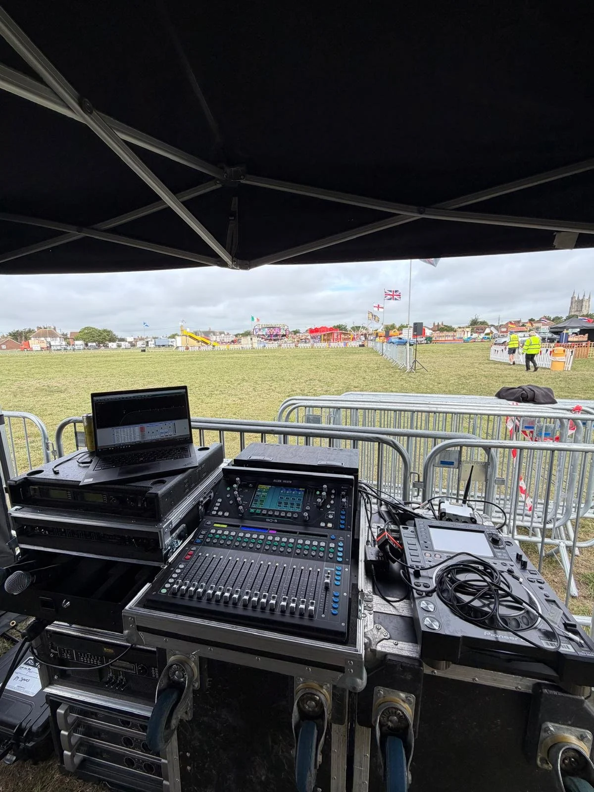 A DJ setup with mixing equipment, laptop, and speakers under a tent at an outdoor fairground. In the background, there are flags, people in high-visibility vests, and fair rides across a grassy field.