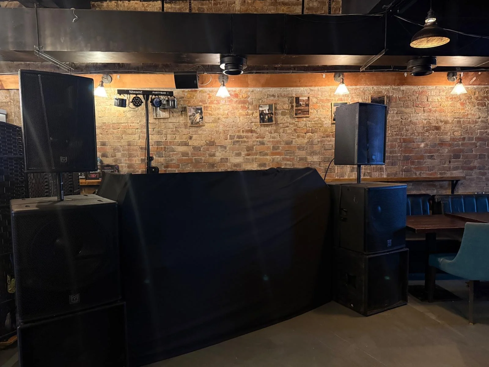 An indoor music venue or bar with large black speakers, a brick wall with small pictures, and stage lighting. There are tables and chairs to the right.