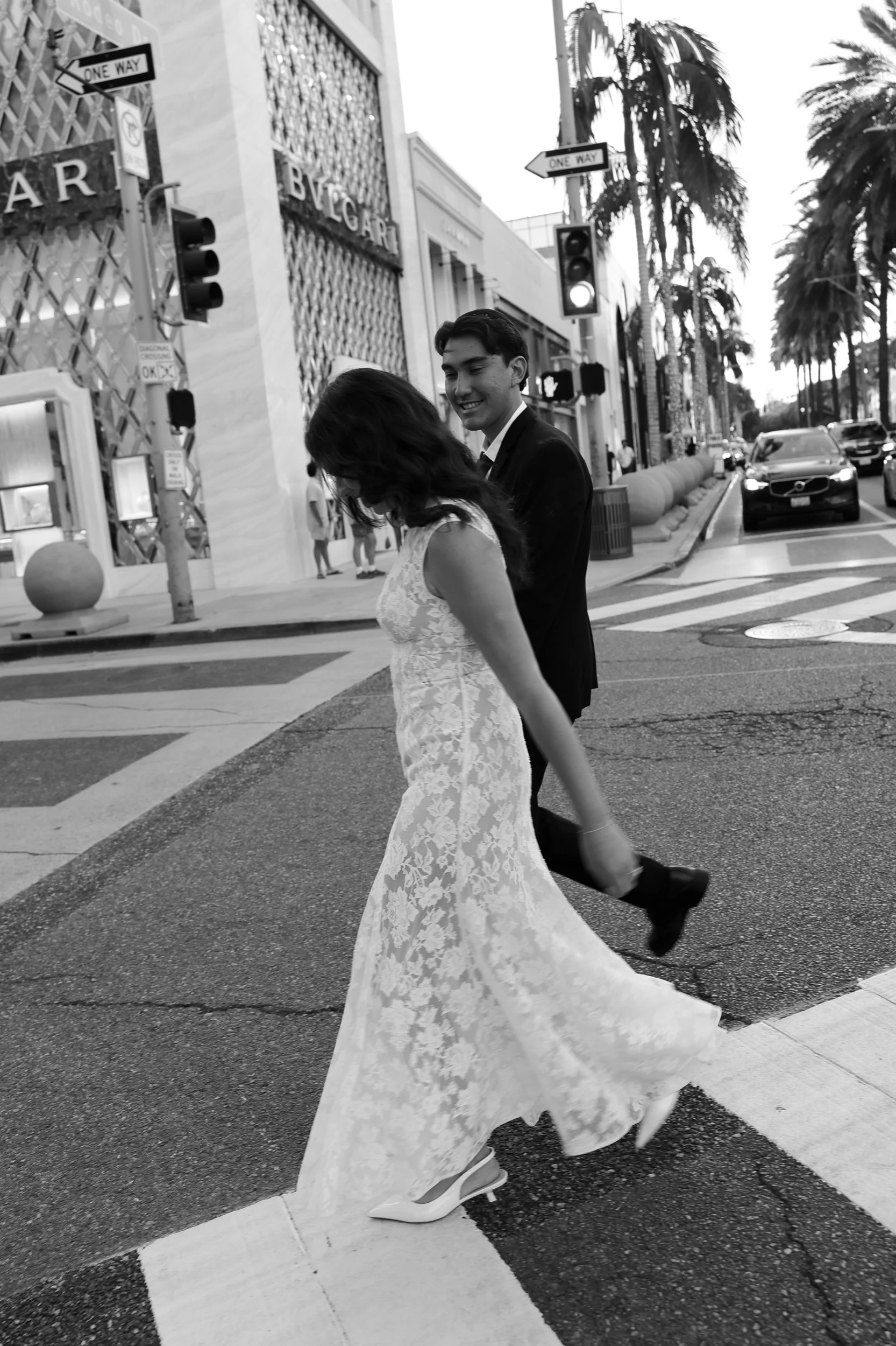 Black-and-white photograph of a bride and groom walking together across a palm-lined street.