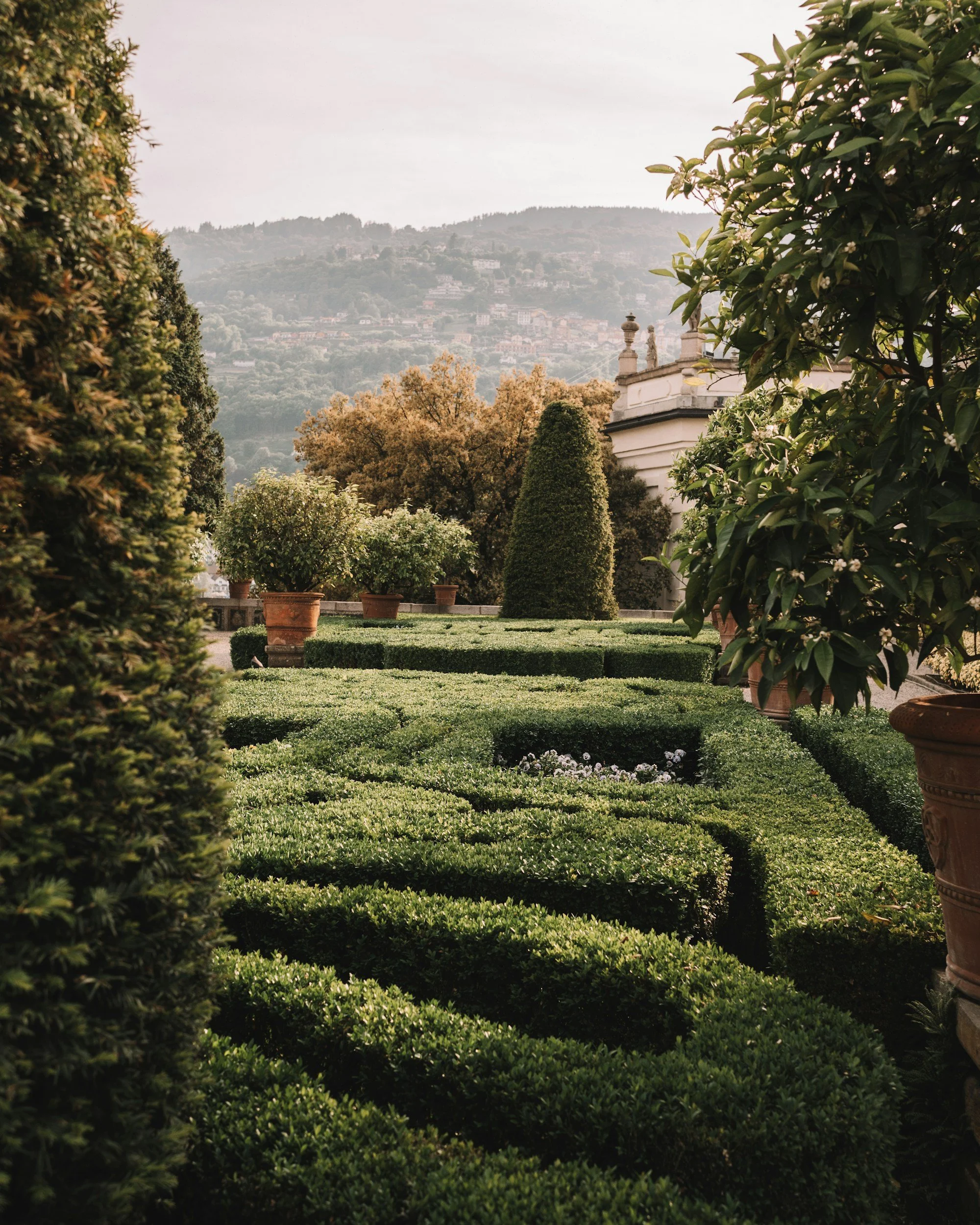 A lush garden with manicured hedges in the foreground, large potted plants, and a hillside with countless buildings in the background, under a cloudy sky.