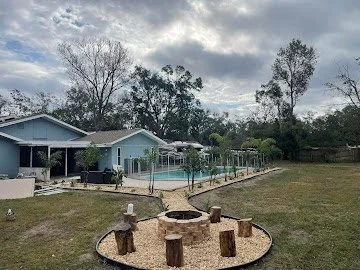 Backyard with a house, swimming pool, and trees under cloudy sky.