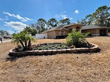 A backyard with a raised flower bed bordered by stones, containing bushes and plants, on a dry lawn with a house and trees in the background under a partly cloudy sky.