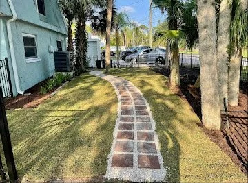 A paved walkway winding through a grassy yard with palm trees, leading to parked cars in a residential area.