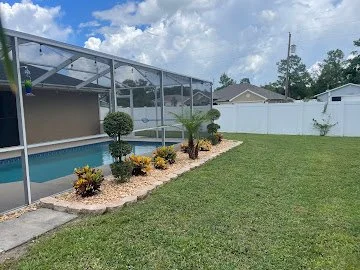 Backyard with a screened-in pool enclosure on the left, a small landscaped area with shrubs and palm trees, a large grassy lawn, and a white fence surrounding the yard under a partly cloudy sky.