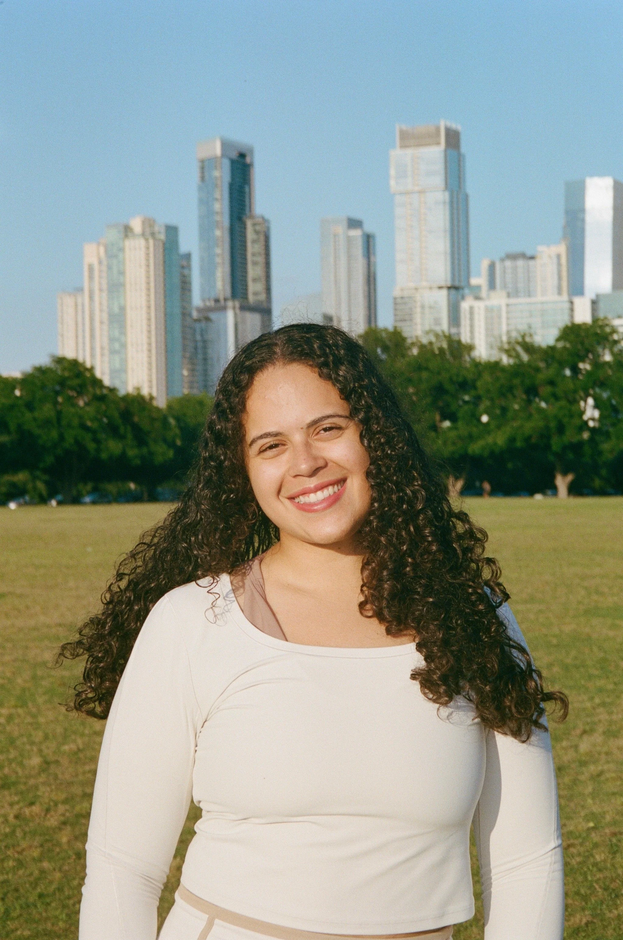 Smiling woman with curly hair standing outdoors in front of a cityscape with tall buildings and green trees, wearing a white long-sleeve top.
