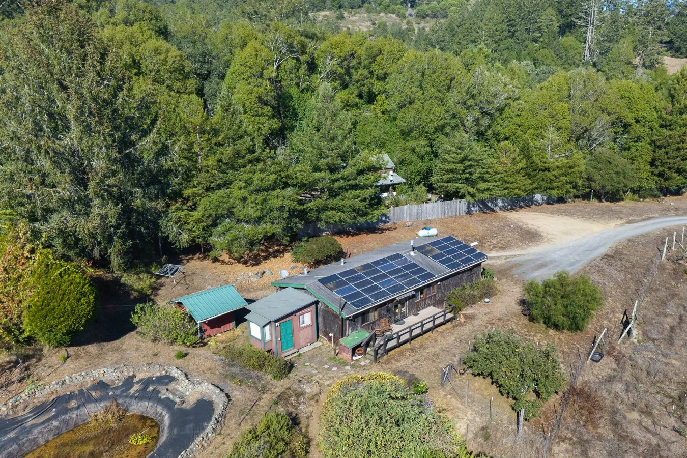 A small house with solar panels on the roof, surrounded by trees and located near a dirt road.