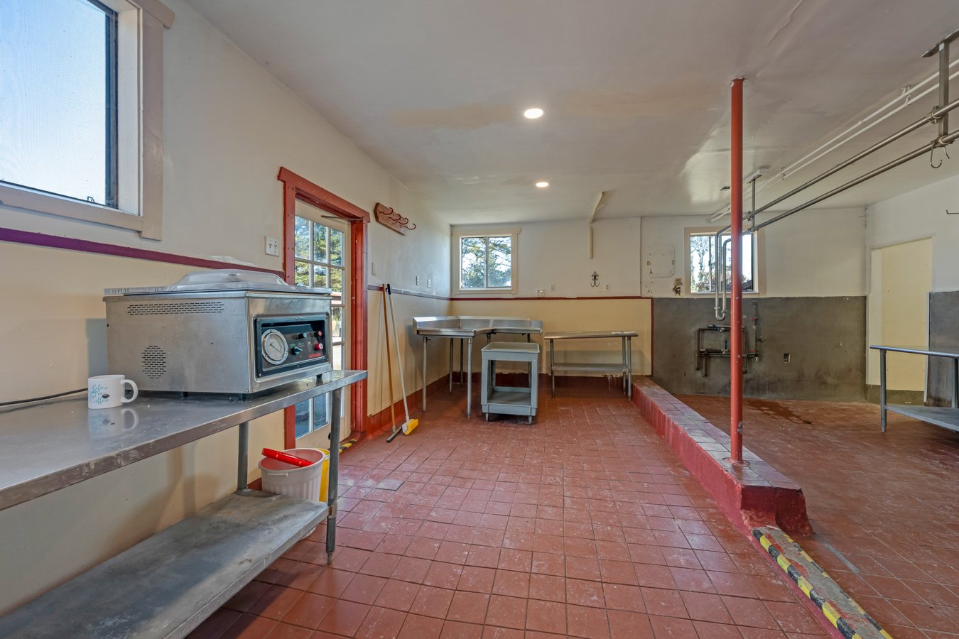A kitchen with red tile flooring, stainless steel counters, and windows. There is a food warmer, cleaning supplies, and empty tables and shelves.