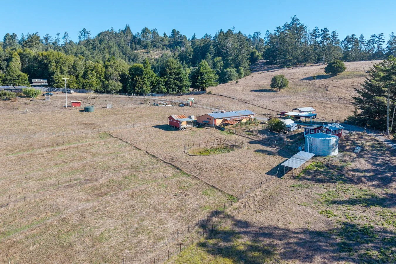 A rural farm property situated in a hilly landscape with several buildings including a large house, sheds, and a water tank, surrounded by dry fields and trees under a clear blue sky.