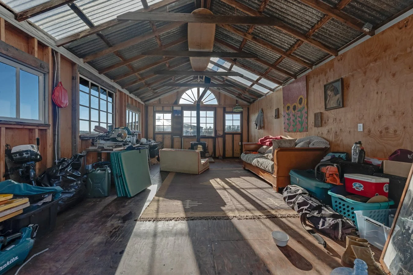 A cluttered rustic shed with wooden walls and a corrugated metal roof, filled with furniture, tools, boxes, and miscellaneous items, illuminated by sunlight coming through multiple windows.