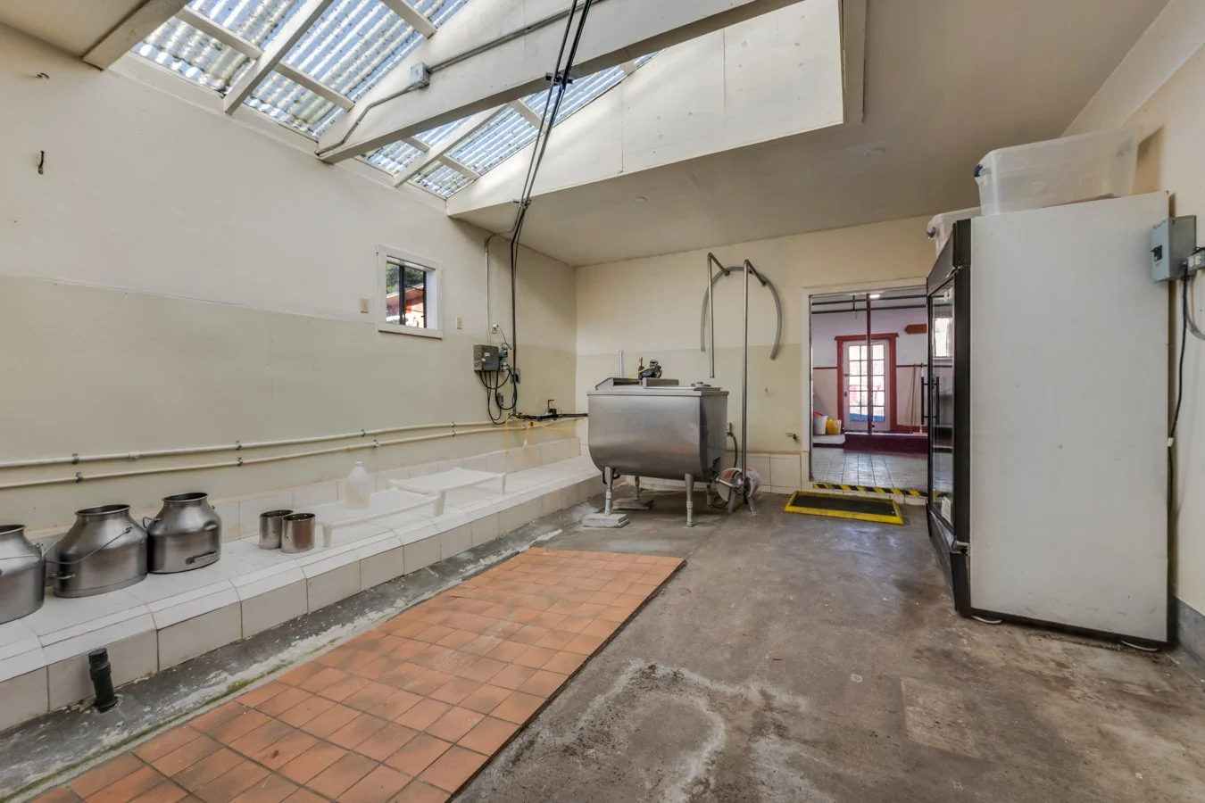 A commercial kitchen or food processing area with tiled surface, stainless steel equipment, a small window, and a skylight ceiling. Walls are white, flooring is concrete with some tiled sections, and there is a doorway leading to another room.