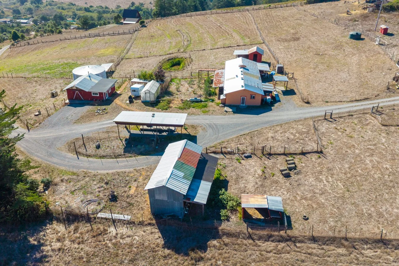 Aerial view of a rural farm with multiple red and gray buildings, fenced fields, and a dirt road running through the property.