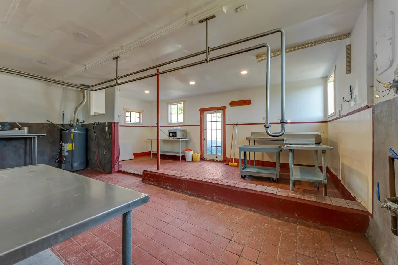 A commercial kitchen under renovation with stainless steel tables, a water heater, cleaning tools, and exposed wiring, featuring a raised platform area with brick flooring and a door leading outside.