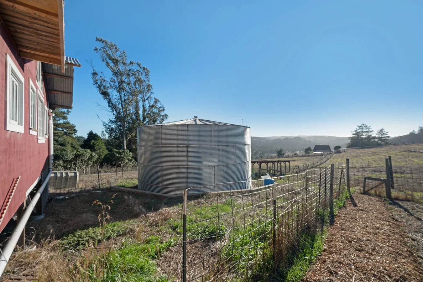 A rural farm scene with a red house on the left, a large metal water tank in the center, and open fields with trees and distant hills under a clear blue sky.