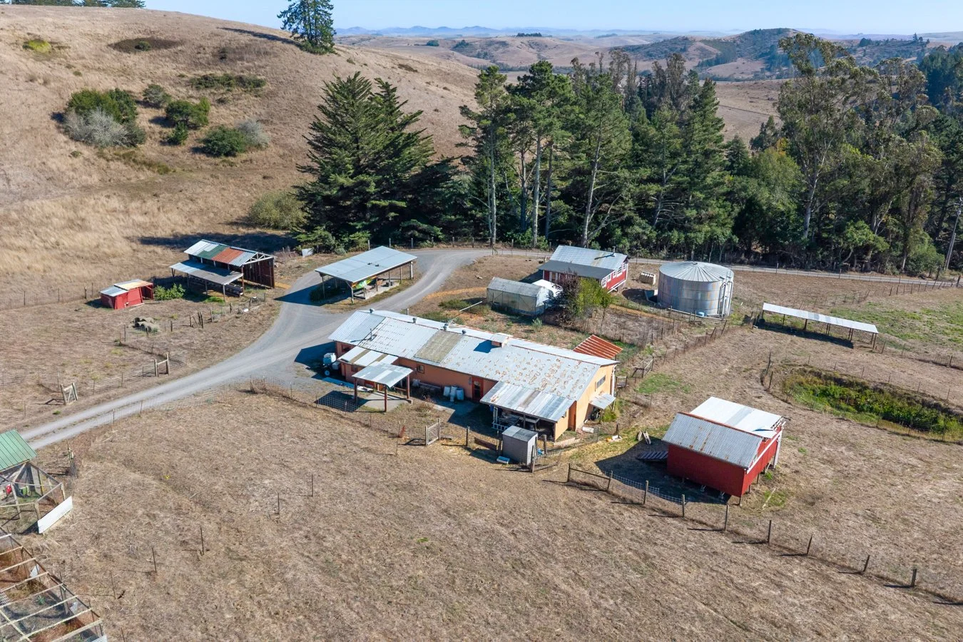 An aerial view of a farm with multiple buildings, farm equipment, and a water tank, surrounded by open fields and distant hills.