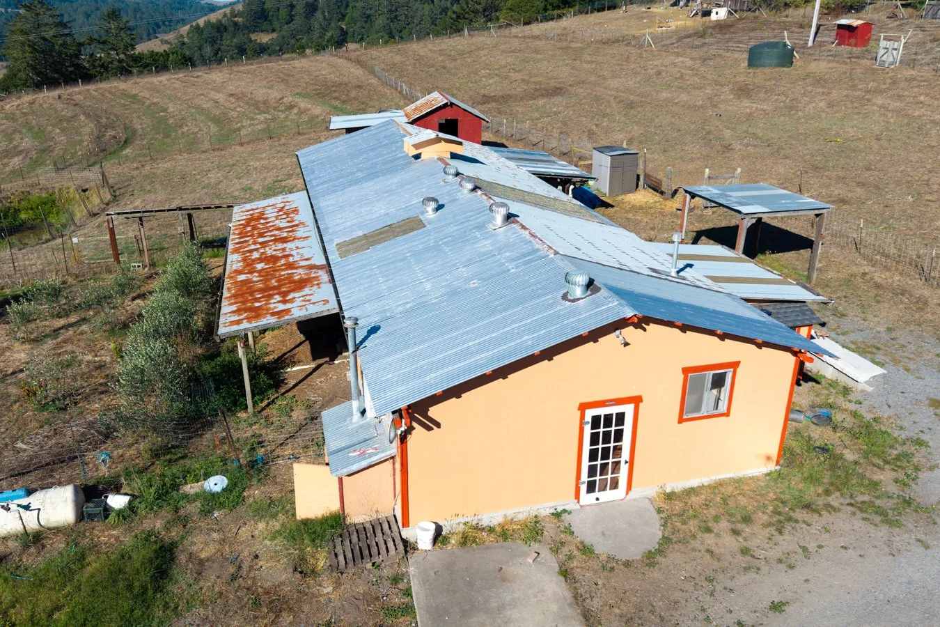 An aerial view of a rural farm building with a metal roof, some parts rusted, and a peach-colored exterior with orange trim. The farm is surrounded by fenced land, with a small red barn and other structures in the background. The terrain is hilly and