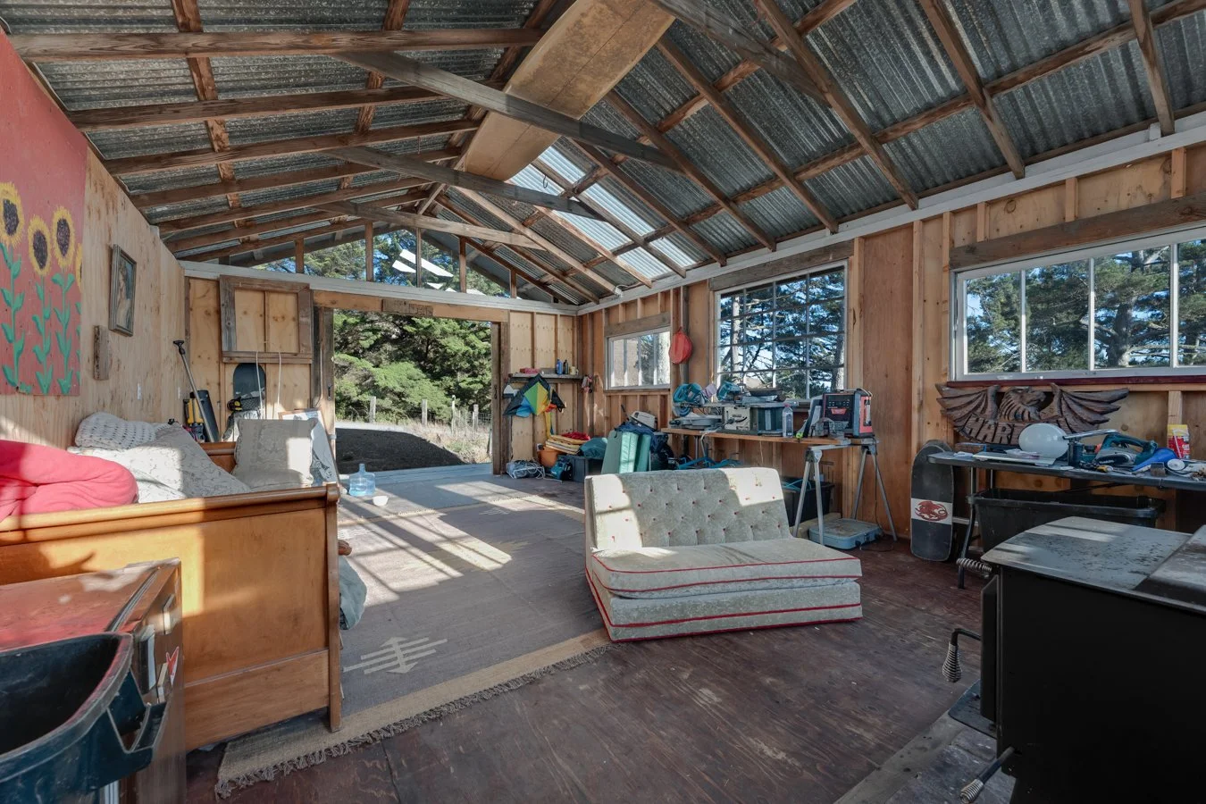Sunlit rustic workshop with wooden walls, a metal roof, and various tools and furniture, including a couch, a vintage chair, and workbenches.
