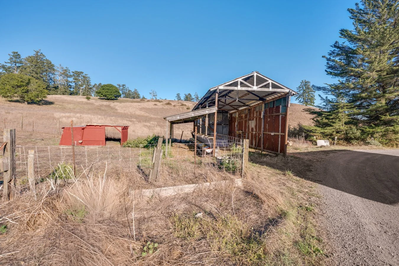 A rural farm scene with a weathered barn with a partially open roof, a small red shed, and a dirt road, surrounded by dry grass, fencing, and trees under a clear blue sky.