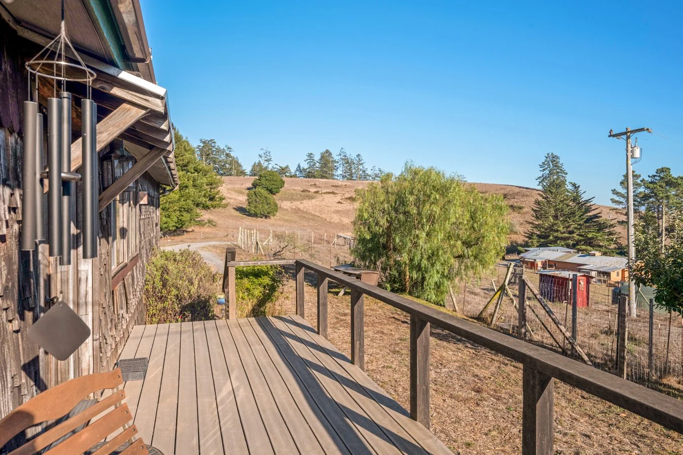 View from a wooden porch with a wind chime, overlooking a rural landscape with trees, hills, and a wire fence, with several small buildings or sheds, a utility pole, and clear blue sky.
