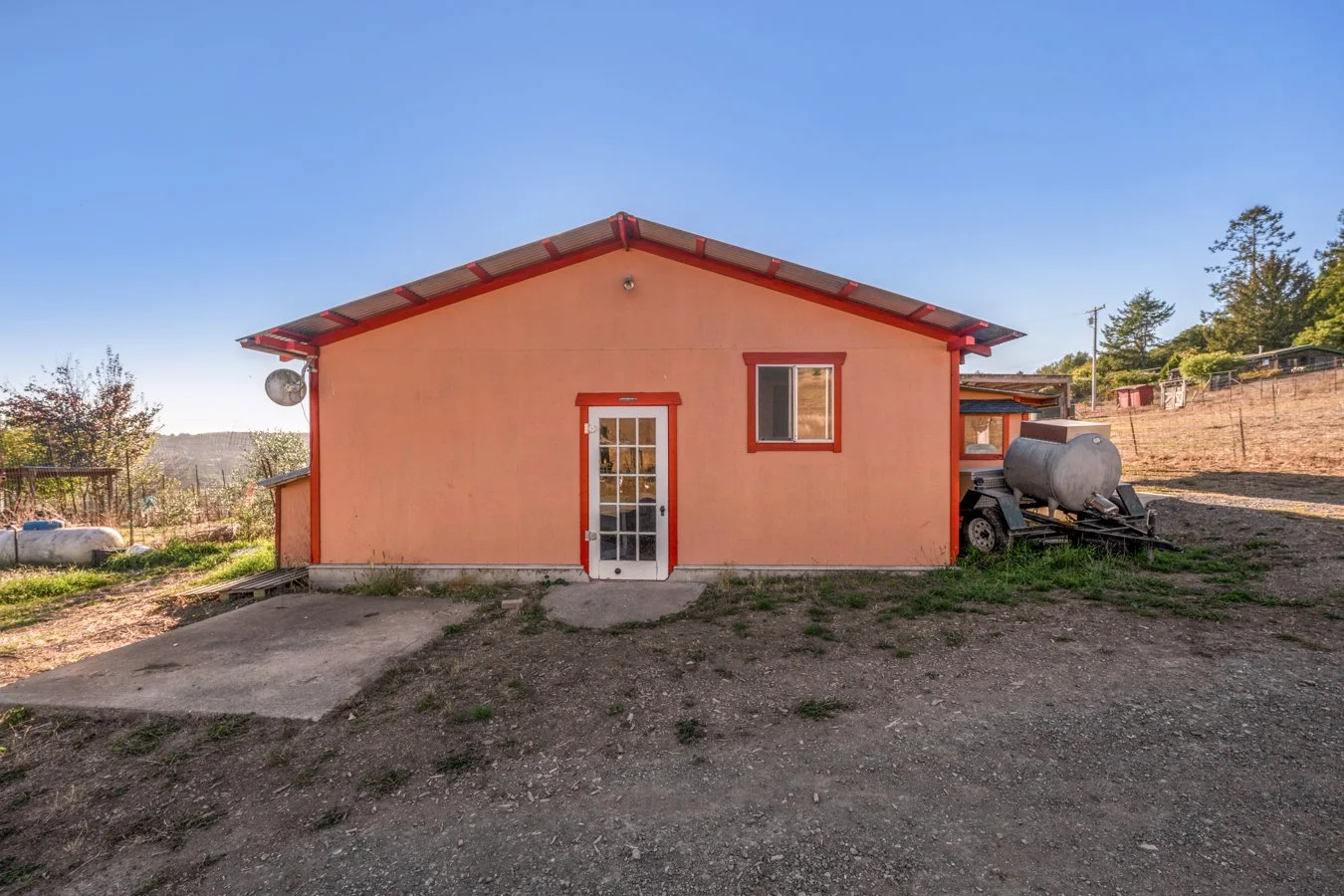 A small peach-colored house with red trim, a glass-paneled door, and a window, set against a rural landscape with dirt ground and distant trees.