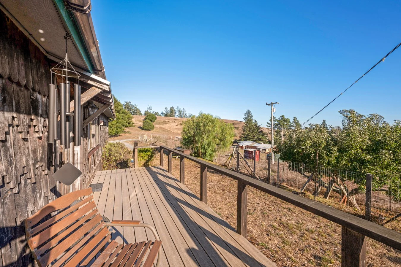 Deck with wooden bench and wind chime, overlooking a rural landscape with trees, fencing, and utility pole under a clear blue sky.
