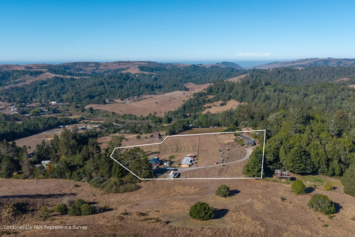 Aerial view of a hillside property outlined in white, with scattered buildings and trees, surrounded by rolling hills and forested areas, with the Sonoma Coast visible in the distance.