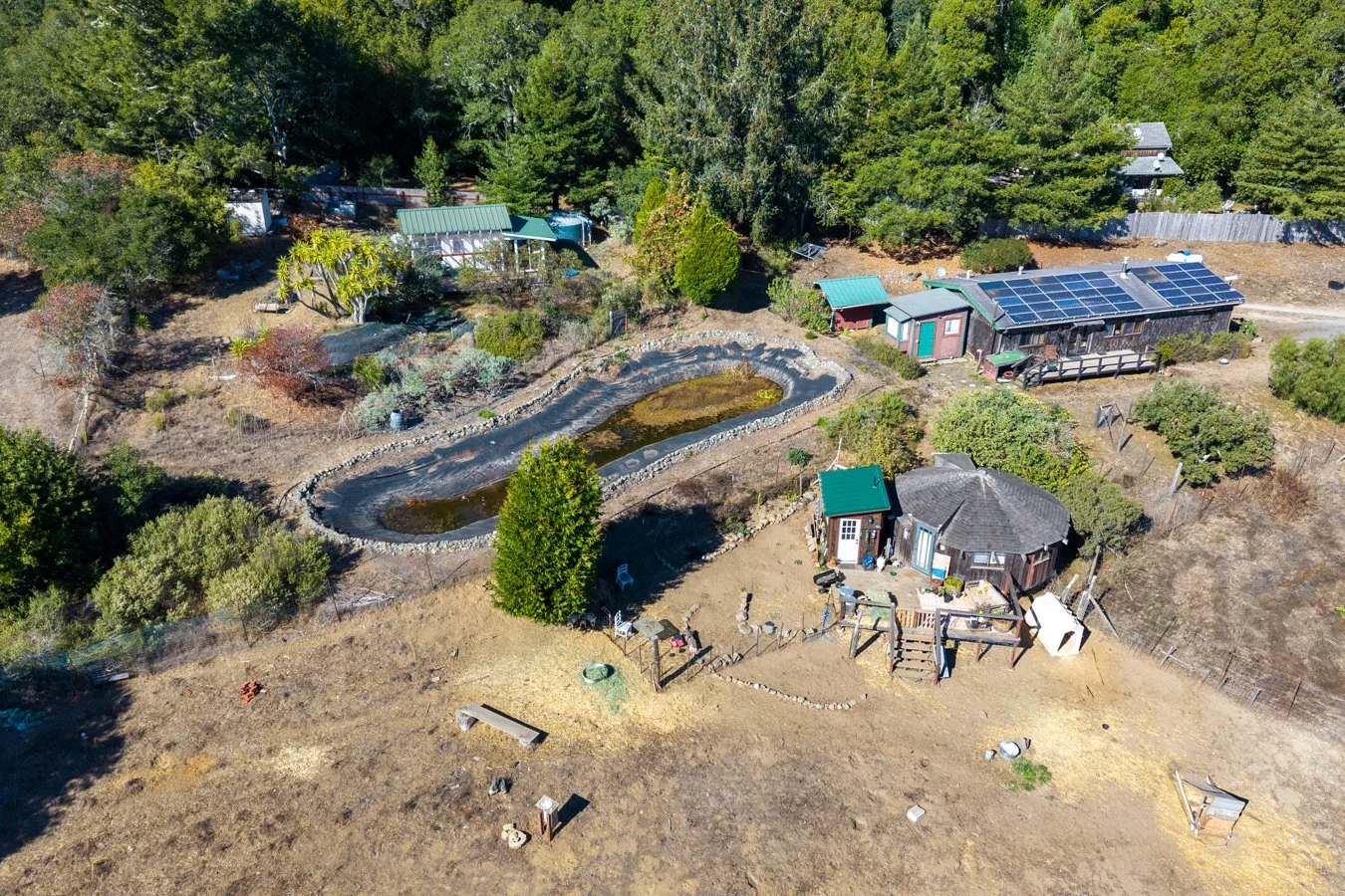 Aerial view of a backyard with a pond, trees, and various small structures, including a house with solar panels on the roof, surrounded by a fenced area and dry grass.