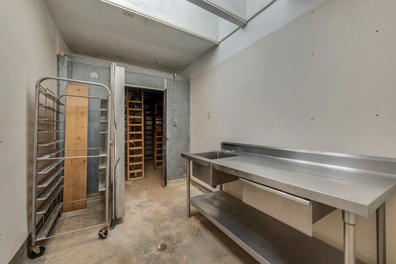 Empty commercial kitchen area with stainless steel sink and shelving, laundry cart, and open doorway leading to storage room with wine racks.