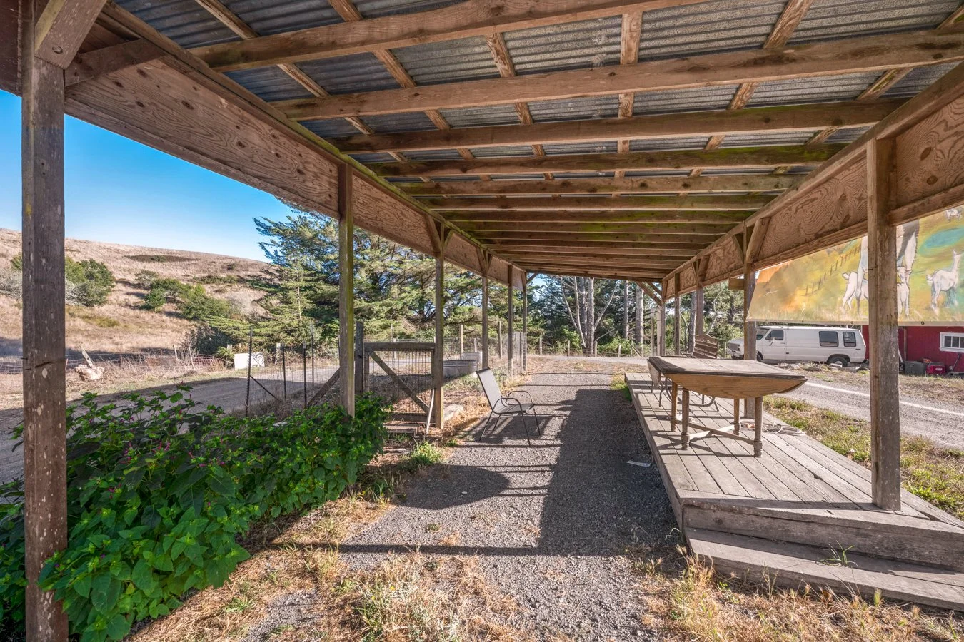 An outdoor patio area with a wooden roof and deck, surrounded by green plants and trees, with a bench and a table under the roof, and a van parked in the background.