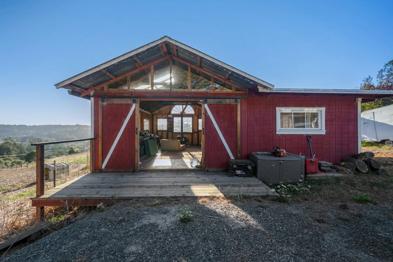 A rural red barn with a wooden deck and an open doorway revealing a rustic interior with furniture, set against a scenic countryside landscape under a clear blue sky.
