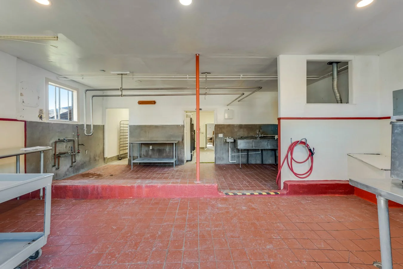 Empty industrial kitchen with stainless steel tables and a sink, red tiled floor, and white walls.