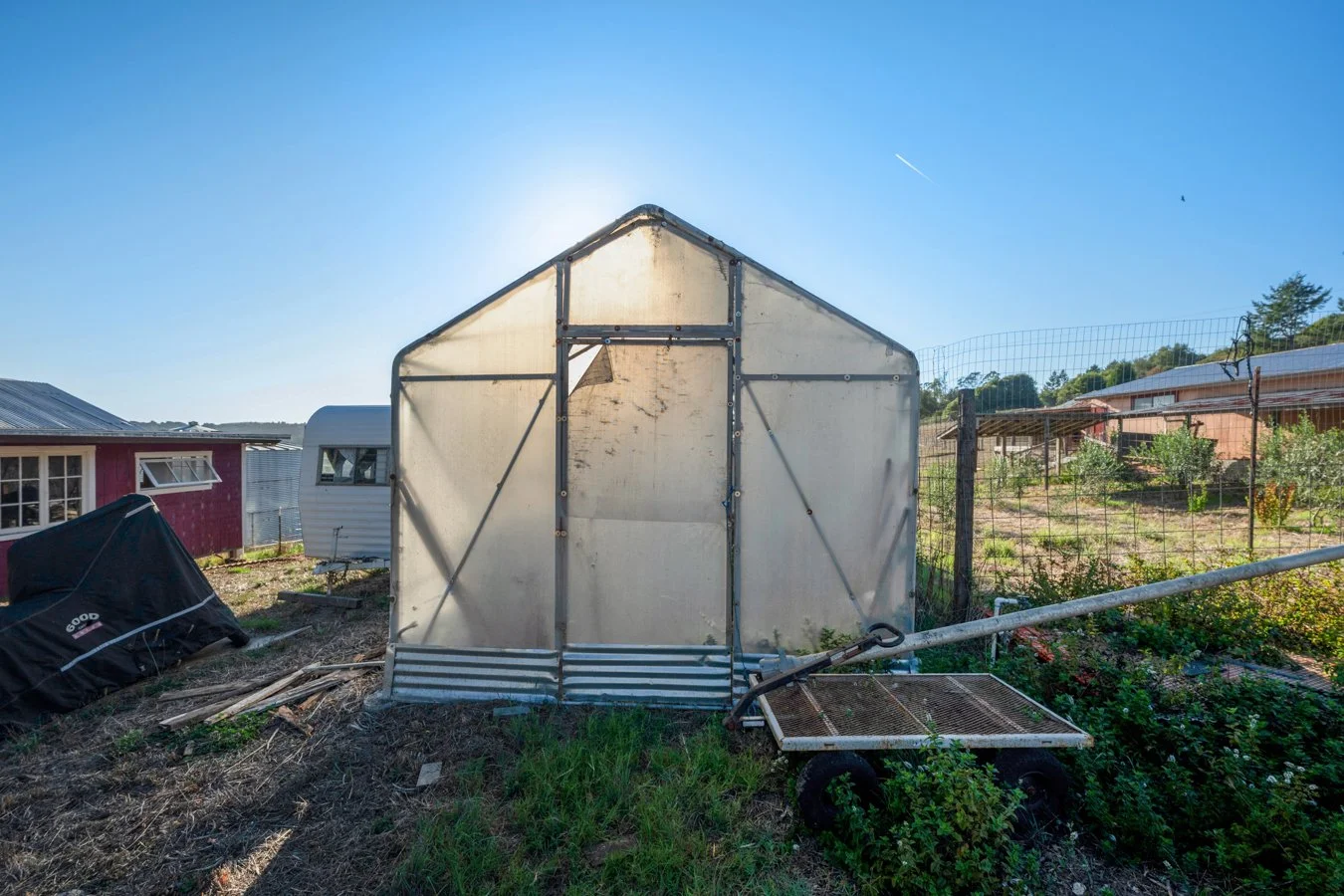 A small greenhouse on a farm with a blue sky, nearby garden, and a wagon with wheels.