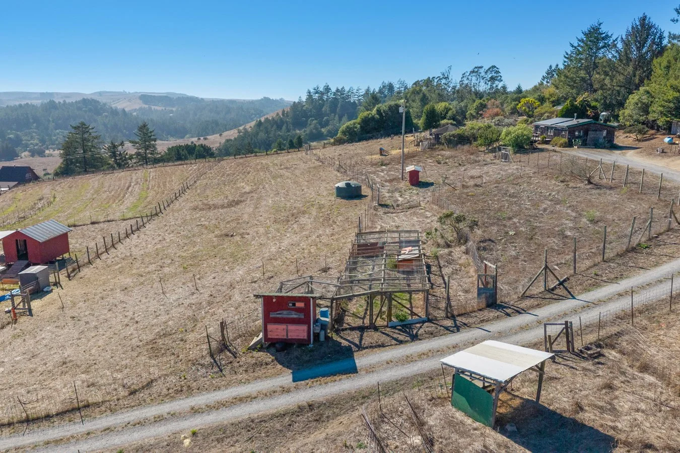 Aerial view of a dry, fenced rural hillside with small structures, grassy patches, dirt paths, trees, and distant hills under a clear blue sky.